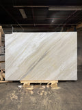 A honed finish Ajax Gold marble slab displayed on a wooden pallet, with a white background featuring diagonal gold, grey, and blue veining.